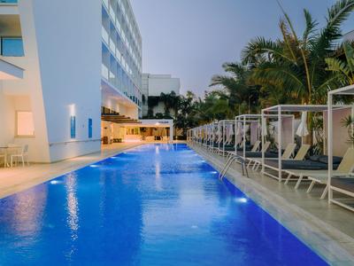 Moderner Hotelpool mit blauer Beleuchtung und Liegestühlen neben einer weißen Gebäudefront bei Dämmerung.
