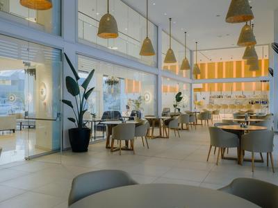 Moderne Hotel-Lounge mit großen Fenstern, runden Tischen und grauen Stühlen in hellem Interieur.
