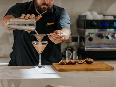 Barkeeper gießt einen Cocktail in ein Glas neben einer Holzplatte mit Orangen.