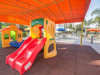 Bunte Kunststoffrutsche mit Überdachung auf Spielplatz, Gummimatte und Palmen im Hintergrund.