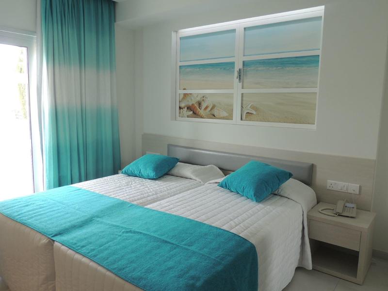 Hell eingerichtetes Schlafzimmer mit Doppelbett, türkisfarbenen Akzenten und Strandbild an der Wand.