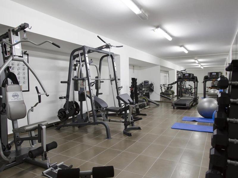 Fitnessraum mit verschiedenen Geräten, Hanteln und Gymnastikmatten, heller Raum mit Fliesenboden.