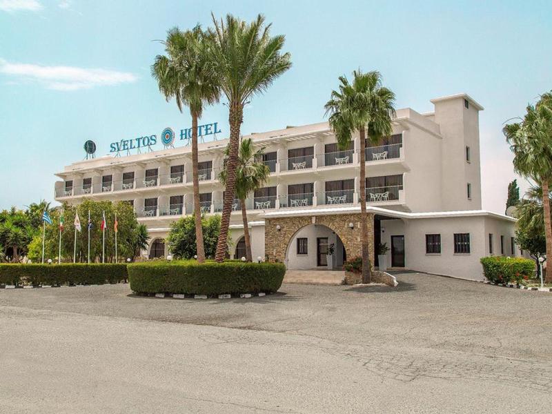 Dreistöckiges Hotelgebäude mit weißen Wänden, Palmen und breitem Hof unter blauem Himmel.