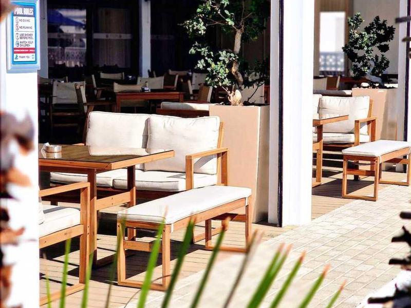 Sonnige Terrasse mit Holzmöbeln, weißen Polstern und grünen Pflanzen in Töpfen.