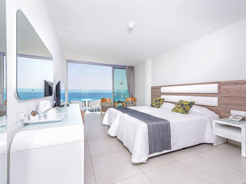 Helles Hotelzimmer mit Doppelbett, Meerblick und Balkon mit Stühlen.