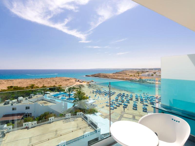 Balkon mit Glasgeländer bietet Meerblick und Poolanlage an einem sonnigen Strandhotel.