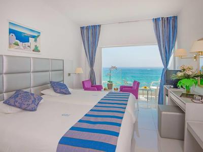 Habitación de hotel luminosa con cama doble, acentos azules y vista al mar desde el balcón.