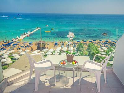 Balcón con sillas blancas y mesa con vista al mar, playa y sombrillas bajo un cielo despejado.