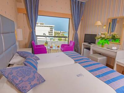 Habitación de hotel luminosa con dos camas, sillones morados y balcón con vista a la ciudad.