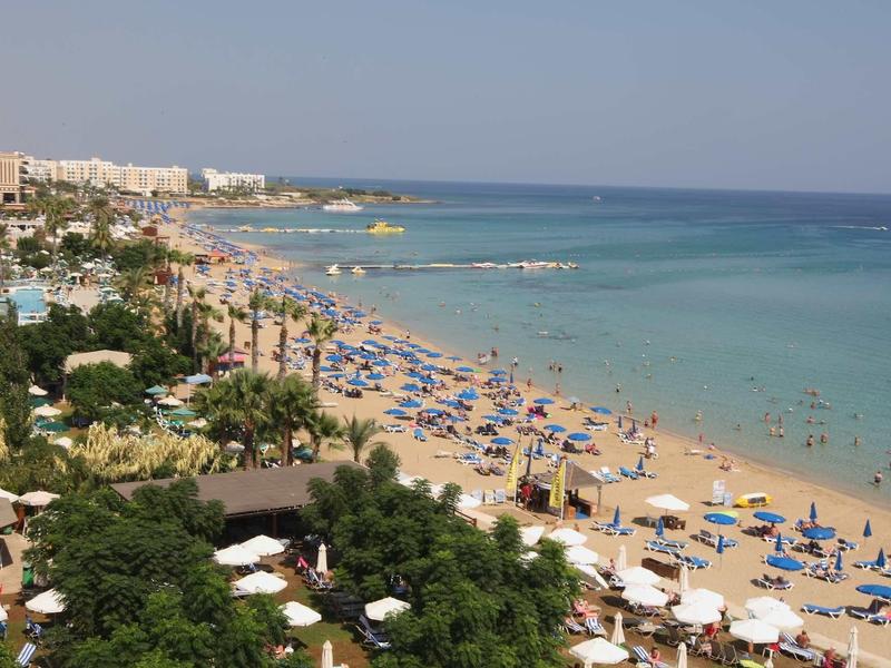 Überfüllter Strand mit blau-gelben Sonnenschirmen, vielen Menschen im Wasser und Gebäuden im Hintergrund.