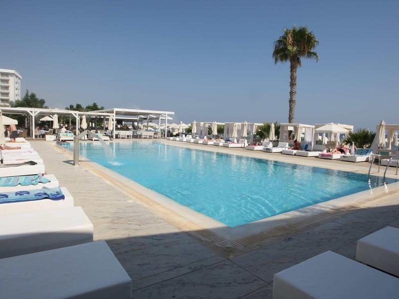 Großer Pool mit blauer Wasserfläche, Liegen und Schirmen unter klarem Himmel und Palmen.