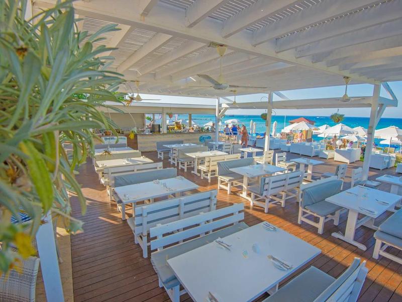 Offenes Strandrestaurant mit weißen Holztischen, blauen Sitzbänken und Blick aufs Meer bei sonnigem Wetter.
