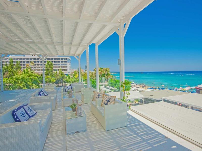 Große Terrasse mit weißen Möbeln, Blick auf Palmen, Hotel und türkisfarbenes Meer bei klarem Himmel.