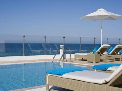Hotelpool mit Liegestühlen und Sonnenschirm am Meer an einem sonnigen Tag.