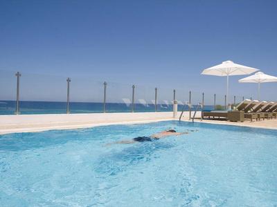 Schwimmbad mit klarem Wasser, Liegestühlen und Sonnenschirmen am Meer unter blauem Himmel.