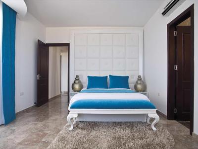 Modernes Hotelzimmer mit weißem Bett, türkisfarbenen Kissen und Gardinen, zwei Nachttischen und Teppichboden.