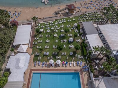 Grüner Garten mit Sonnenschirmen und Liegen neben blauem Schwimmbecken und Strand im Hintergrund.