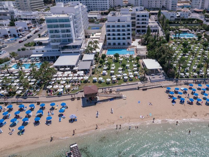 Strand mit blauen Sonnenschirmen, Sand und Meer, dahinter Hotels und Palmen in Reihen.