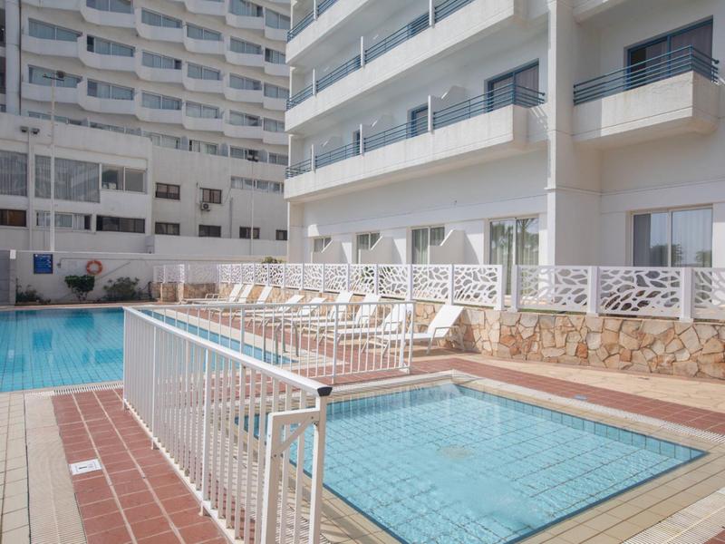 Mehrstöckiges Gebäude mit Balkon, Pool und Kinderbecken im Vordergrund, weiße Liegestühle.