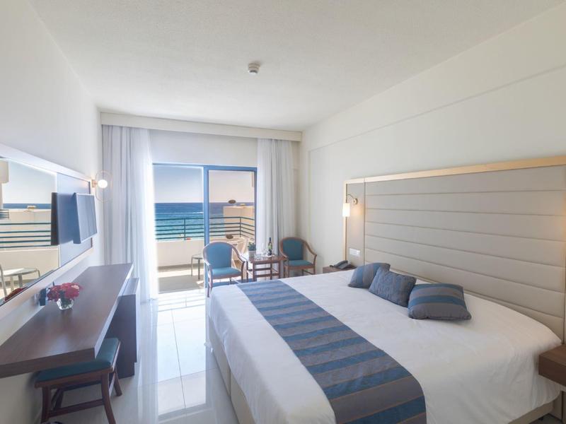 Helles Hotelzimmer mit großem Bett, Schreibtisch, Balkon und Meerblick bei Tageslicht.