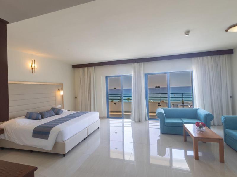 Helles Hotelzimmer mit Doppelbett, blauen Sofas, großem Fenster und Meerblick.