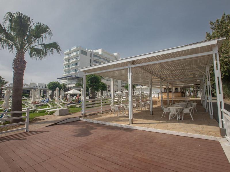 Moderner Außenbereich mit Holzterrasse, überdachtem Sitzbereich, Palmen und Hotelgebäude im Hintergrund.