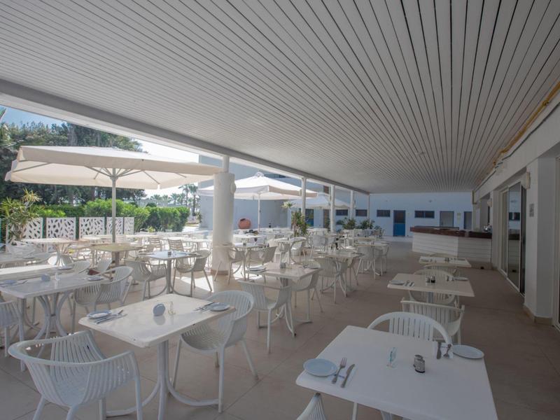 Große überdachte Terrasse mit weißen Tischen, Stühlen und Sonnenschirmen an sonnigem Tag.