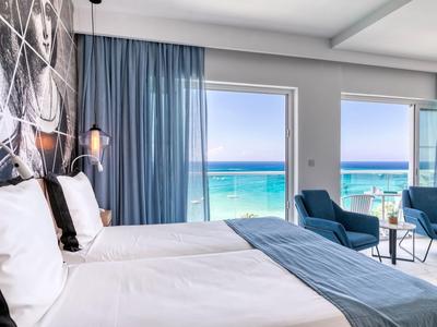 Chambre d'hôtel avec deux lits et vue sur la mer à travers de grandes fenêtres avec ciel bleu.