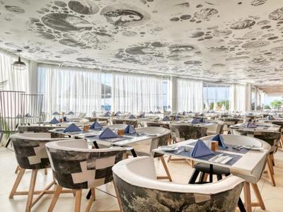 Ristorante luminoso e raffinato con tavoli rotondi, sedie decorate e grandi finestre.