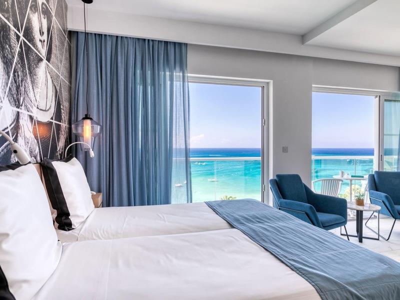 Chambre d'hôtel avec deux lits et vue sur la mer à travers de grandes fenêtres avec ciel bleu.