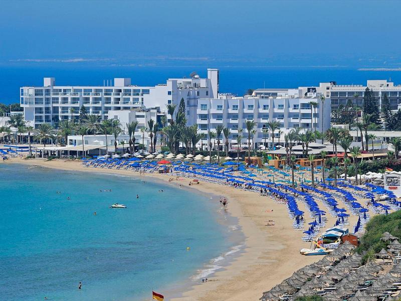 Strand mit blauem Wasser und einer Reihe von blauen Sonnenschirmen vor einem großen Hotelgebäude.