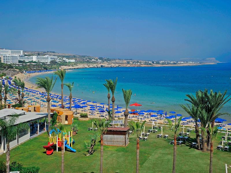 Strand mit blauem Meer, Sonnenschirmen und Liegestühlen, im Vordergrund ein Spielplatz und Palmen.