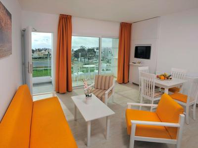 Helles Hotelzimmer mit orangem Sofa, Sesseln und Blick auf Grünfläche durch große Fenster.