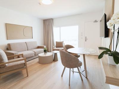 Modern eingerichtetes Hotelzimmer mit Sofa, Sesseln und Essbereich bei Tageslicht.