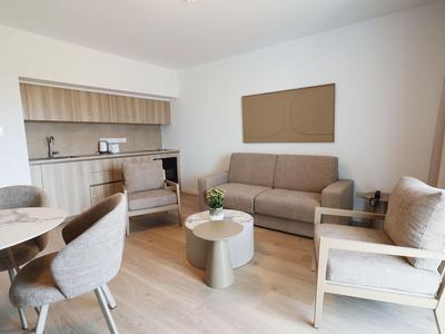 Modern eingerichtetes Hotelzimmer mit Sofa, Stühlen, Tisch und Küchenzeile in hellen Farben.