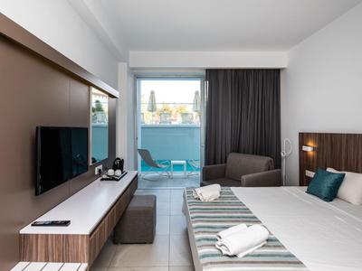 Helles Hotelzimmer mit Doppelbett, Balkon, Sessel und Flachbildfernseher.