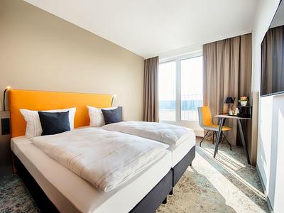 Chambre d'hôtel lumineuse avec lit double, tête de lit jaune, bureau et grande fenêtre.