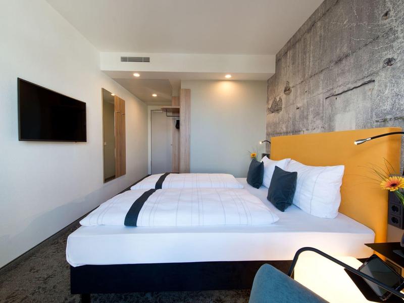 Chambre d'hôtel moderne avec un grand lit, une tête de lit jaune, une TV murale et une décoration neutre.