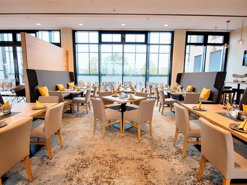 Grande salle à manger d'hôtel avec chaises beiges, tables en bois et grandes fenêtres laissant entrer la lumière naturelle.