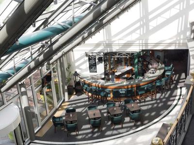 Brightly lit hotel lounge with curved bar area and rows of small tables and chairs.