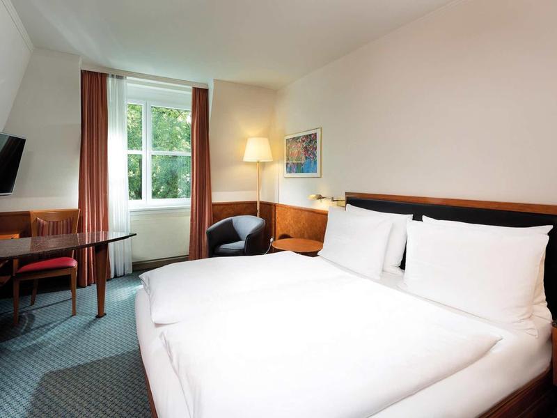 Bright hotel room with large bed, desk, armchair, and window view of greenery.