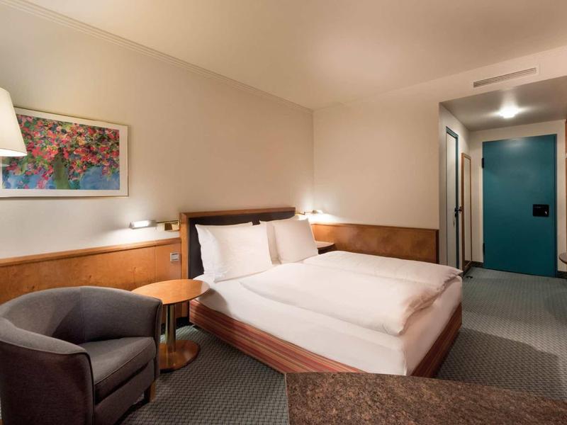 Modern hotel room with large double bed, armchair, bedside table, and ambient lighting.