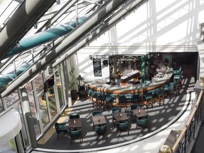 Brightly lit hotel lounge with curved bar area and rows of small tables and chairs.