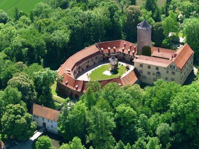 Luftaufnahme einer historischen Burg mit halbkreisförmigem Gebäudeteil, umgeben von dichtem Wald.