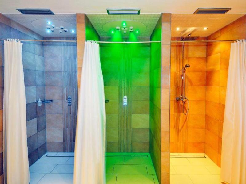 Tři moderní sprchy s bílými závěsy a barevným osvětlením v hotelové koupelně.