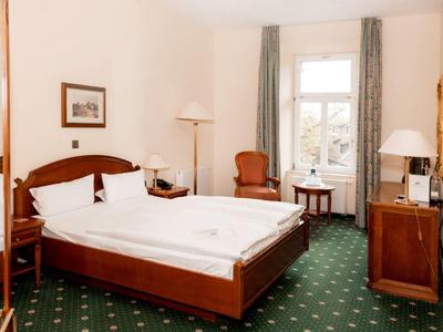 Helles Hotelzimmer mit Doppelbett, grünem Teppich und zwei Sesseln am Fenster.