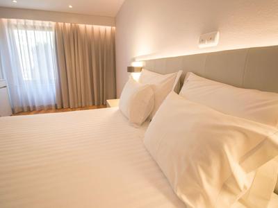 Chambre d'hôtel lumineuse avec grand lit, oreillers blancs et rideaux à la fenêtre.