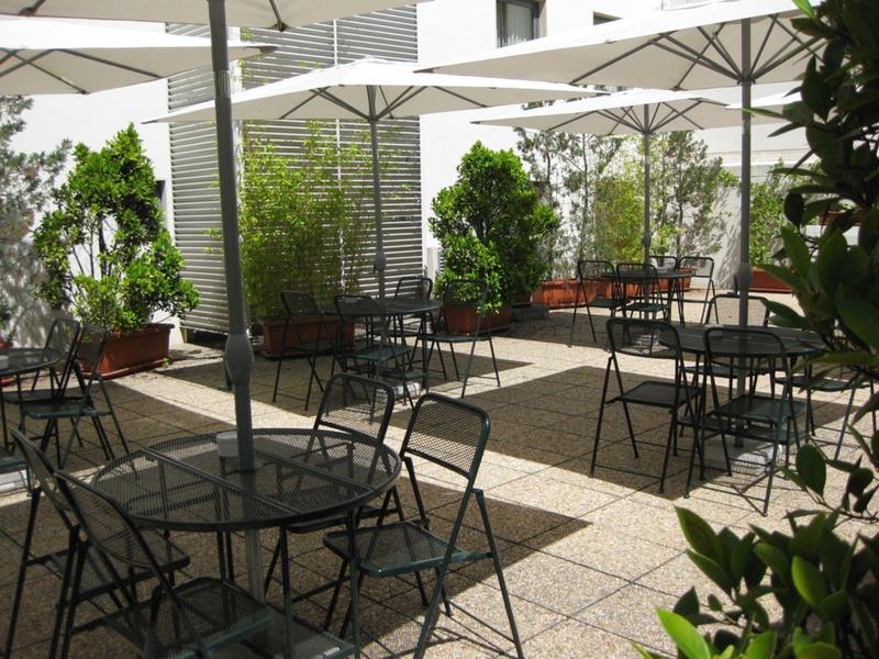 Espace extérieur ensoleillé avec tables, chaises et parasols à côté d'un bâtiment hôtelier.