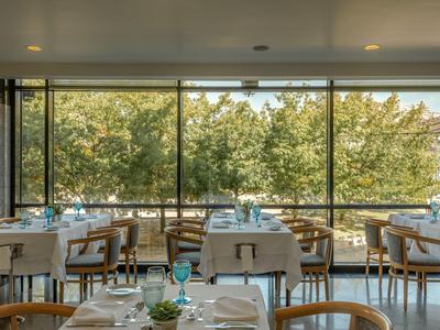 Licht restaurant met houten stoelen, witte tafelkleden en grote ramen met uitzicht op groene bomen.