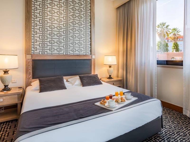 Licht ingerichte hotelkamer met groot bed, nachtkastjes en een ontbijtplankje.